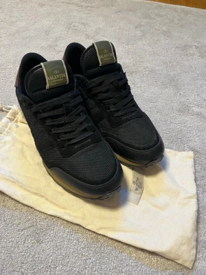 Svarta Valentino Garavani sneakers - Snygga svarta sneakers från Valentino Garavani med mesh och mockadetaljer. Skorna har svarta snören, gröna logotungor och en klassisk låg siluett. Perfekta för dig som vill ha en stilren och exklusiv look.