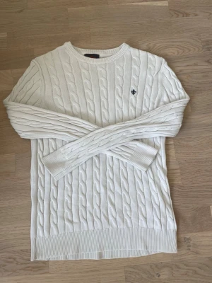 Vit kabelstickad tröja - Kabelstickad vit tröja med rund halsringning och ribbade muddar. Tröjan har ett diskret broderat emblem på bröstet och är tillverkad i ett mjukt material som passar perfekt för kyligare dagar. Klassisk och stilren design med lång ärm. Hör av er vid frågor eller prisförslag🤗
