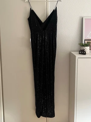 Svart långklänning med paljetter - Snygg svart långklänning med tunna axelband och omlott-ringning. Klänningen är täckt av glittriga paljetter som ger en festlig look. Perfekt för dig som vill sticka ut och glänsa på nästa event. Materialet känns mjukt och följsamt.
