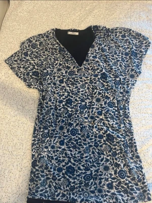 Blommig topp från MNG Suit Collection - Snygg topp från MNG Suit Collection med blått och vitt blommönster. Toppen har v-ringning och korta ärmar, perfekt för dig som gillar en avslappnad men ändå stilren look. Materialet känns mjukt och lätt, vilket gör den skön att bära.