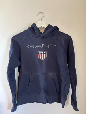 Mörkblå hoodie från GANT - Mörkblå hoodie från GANT med broderad logga och amerikanskinspirerat sköldmärke på bröstet. Klassisk känguruficka framtill och huva. Tillverkad i mjukt bomullsmaterial, perfekt för en avslappnad och stilren look.