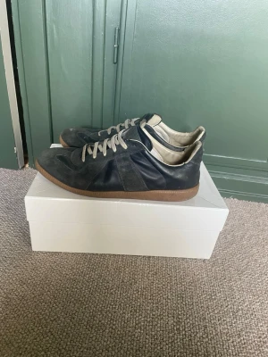 Maison margiela gats - Säljer ett par svarta sneakers med vita skosnören och bruna sulor. Skorna är låga och har en snygg mix av mocka och läder på ovandelen. Insidan är ljus och de har en klassisk, stilren look som passar till det mesta.