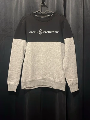 Grå och svart Sail Racing sweatshirt - Snygg sweatshirt från Sail Racing i tvåfärgad design med svart upptill och ljusgrå nedtill. Tröjan har rund hals, ribbade muddar och logga tryckt på bröstet. Perfekt för en avslappnad och sportig stil.