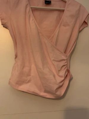 Ljusrosa omlottopp från Gina Tricot - Supersöt ljusrosa omlottopp från Gina Tricot i mjuk bomull. Toppen har korta ärmar, v-ringning och snygg rynkdetalj i sidan. Perfekt att styla med jeans eller kjol för en fräsch look.