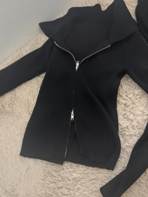 Svart ribbad ziptröja med hög krage - Snygg svart ribbad tröja med dragkedja framtill och hög krage. Tröjan har långa ärmar och är tillverkad i ett stretchigt material som sitter skönt på kroppen. Perfekt för lager på lager och enkel att matcha med olika outfits.