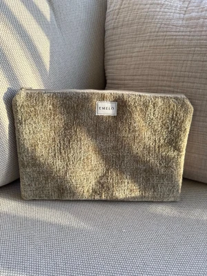 Beige/guld necessär/fodral medium från Emelo - Stilren necessär i beige tyg från Emelo. Väskan har en rektangulär form och dragkedja upptill. Materialet är mjukt och har en lätt textur som ger ett exklusivt intryck. Perfekt för att organisera småsaker i väskan eller på resan. Passar även för iPad. Mått ca 29x20cm