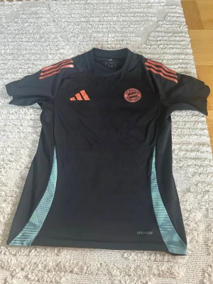 Svart Bayern München träningströja - Säljer en svart träningströja från Adidas med Bayern München-logga på bröstet. Tröjan har korallfärgade detaljer på axlar och loggor samt ljusblå mönstrade paneler på sidorna. Tillverkad i funktionsmaterial med korta ärmar och rund hals. Storleken är XS men passar som S