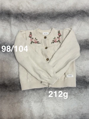  kofta med broderade blommor - Söt kofta med broderade rosa och gröna blommor vid axlarna. Koftan har rund hals, ribbade muddar och tre bruna knappar framtill. Perfekt för lager-på-lager och en mysig stil.🌸 Fint använt skick. Storlek 98/104