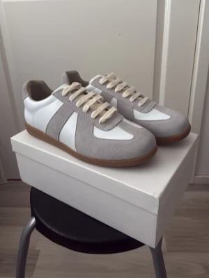 Maison Margiela gråvita 43 - Snygga sneakers i vitt och ljusgrått med ovandel i en mix av slätt läder och mocka. Skorna har beigea snören och en klassisk gummisula i brunt. Perfekta för en clean och stilren look till vardagsoutfiten.