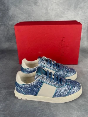 Valentino flycrew - Ett par spektakulära Valentino Garavani Flycrew sneakers helt täckta i gnistrande blått glitter med detaljer i metalliskt blått läder, vit sidorand samt de ikoniska Rockstud-nitarna längs hälen. En perfekt statement-sko för dig som vill kombinera elegans och lyx med en unik streetwear-stil. Skorna säljs i näst intill nyskick, samtliga nitar på baksidan är helt intakta, sulorna visar endast minimalt med spår av användning och innersulorna är rena och fräscha. 