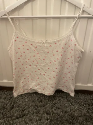 Vit linne med rosa mönster och spets - Sött vitt linne med tunna axelband och små rosa rosetter som mönster. Spetskant upptill och en liten rosett framtill ger en gullig touch. Perfekt till sommaren eller som ett lagerplagg. Mjukt och bekvämt material.