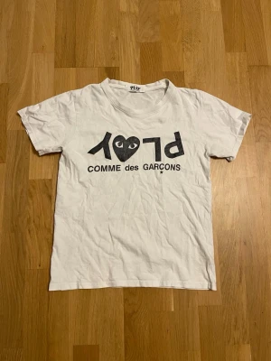Comme des Garçons Play t-shirt - Vit t-shirt från Comme des Garçons Play med svart tryck på framsidan där det står 'PLAY' och ett hjärta med ögon. Klassisk rund hals och korta ärmar. Perfekt för dig som gillar stilrena och ikoniska designer. Texten på tröjan är också lite sprucken.den är storlek S men har krympt i tvätten och är nu mer storlek xs