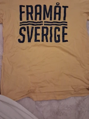 Gul t-shirt med texten FRAMÅT SVERIGE - Gul t-shirt med stor mörkblå text 'FRAMÅT SVERIGE' och grafiska linjer på bröstet. Klassisk rak passform och rund halsringning. Perfekt för dig som vill visa ditt stöd för Sverige på ett stilrent sätt.