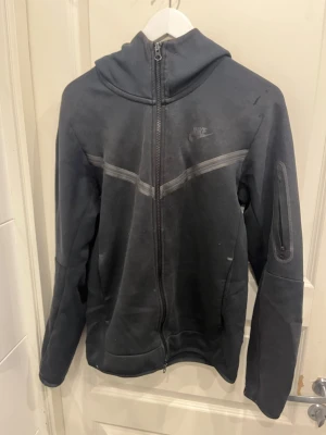 Svart Nike hoodie med dragkedja - Säljer en svart hoodie från Nike med hel dragkedja framtill och huva. Hoodien har en diskret Nike-logga på bröstet, en ficka med dragkedja på ärmen och ett stilrent mönster över bröstet. Perfekt för dig som gillar sportig och avslappnad stil.