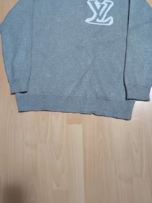 Grå stickad tröja Louis Vuitton - Snygg grå stickad tröja från Louis Vuitton med vit LV-logga på bröstet. Tröjan har ribbstickade muddar vid ärmslut och nederkant. Perfekt för dig som gillar exklusiva och stilrena plagg.