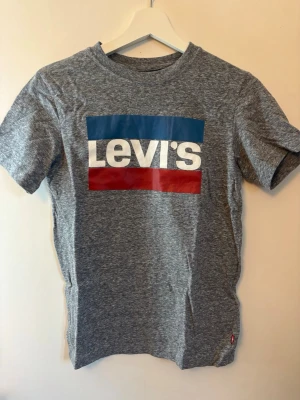 Grå Levi's t-shirt  - Snygg gråmelerad t-shirt från Levi's med klassisk stor logga i blått, vitt och rött framtill. T-shirten har korta ärmar och rund hals. Tillverkad i mjuk bomull för skön känsla och passar perfekt till jeans eller shorts.