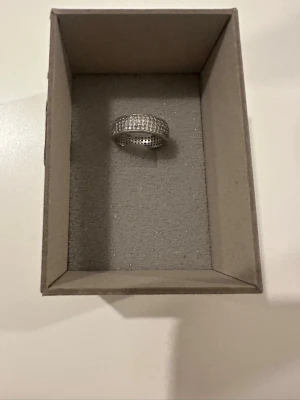 Bred silverfärgad ring med stenar - Säljer en bred och glittrig ring i silverfärg med flera rader små gnistrande stenar runt hela ringen. Ringen har en modern och lyxig känsla och passar perfekt för dig som gillar att sticka ut med dina accessoarer.