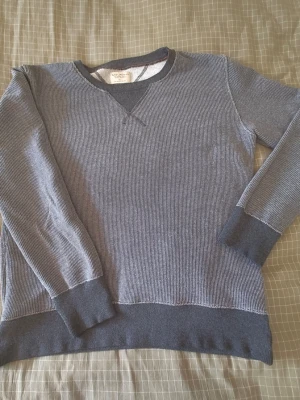 Grå långärmad tröja från Nudie Jeans - Grå långärmad tröja från Nudie Jeans i ribbad bomull. Tröjan har mörkare muddar vid ärmslut och nederkant samt en klassisk rund halsringning med V-detalj framtill. Perfekt för en avslappnad och stilren look.