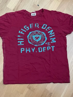 Vinröd Hilfiger Denim t-shirt - Snygg vinröd t-shirt från Hilfiger Denim med stort turkost tryck framtill. Klassisk rund hals och korta ärmar. Perfekt till jeans eller shorts för en avslappnad look.