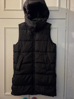 Svart lång väst med huva 134-140  - Snygg svart lång väst med stor huva och dragkedja framtill. Västen har två snedställda fickor och quiltad design som ger en modern look. Perfekt för lager-på-lager och håller dig varm under kyliga dagar. Materialet är mjukt och puffigt.