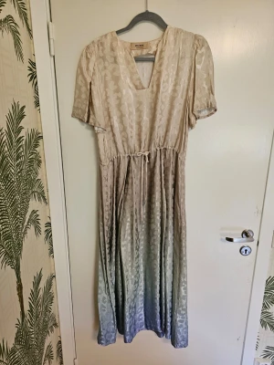 Midiklänning - Grade Dress - Elegant midiklänning i ljusa beige och blå toner. Klänningen har korta ärmar, v-ringning och ett knyt i midjan för en snygg siluett. Materialet har en lätt glansig finish som ger en exklusiv känsla. Använd en gång. Nypris 1 999 kr. 