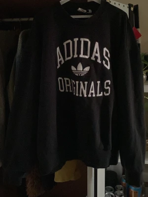 Svart Adidas Originals stickad tröja - Säljer en svart stickad tröja från Adidas Originals med vit text och logga framtill. Tröjan har rund halsringning och långa ärmar, perfekt för en avslappnad streetwear-look. Klassisk och enkel design som passar till det mesta.