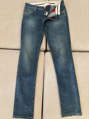 Blå raka jeans från Tramarossa - Säljer ett par blå jeans från Tramarossa med raka ben och klassisk femficksmodell. Jeansen har snygga slitningar och kontrastsömmar i rött samt knappgylf med märkets logga. Materialet är mjukt denim som sitter skönt hela dagen.