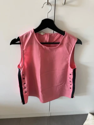 Rosa ärmlös linnetopp med knappar - Snygg rosa ärmlös linnetopp med svarta detaljer längs sidorna och dekorativa knappar. Toppen har rund halsringning och är gjord i ett glansigt material som känns mjukt mot huden.