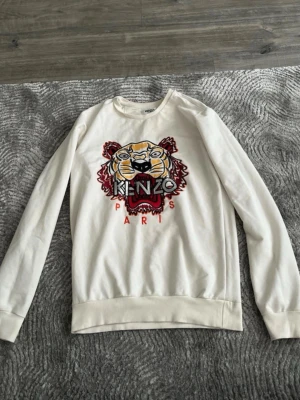 Vit sweatshirt med tigertryck från Kenzo - Säljer en vit sweatshirt från Kenzo med det ikoniska tigerhuvudet broderat framtill och texten 'KENZO PARIS' i rött och svart. Tröjan har rund halsringning, långa ärmar och ribbade muddar. Perfekt för dig som gillar streetwear och vill sticka ut.