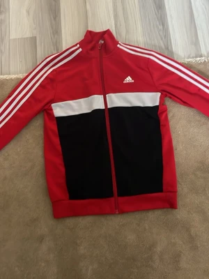 Röd Adidas track jacket med vita ränder - Snygg röd Adidas track jacket med klassiska vita ränder längs ärmarna och vit logga på bröstet. Jackan har svart panel framtill och vit detalj över bröstet. Dragkedja hela vägen och hög krage. Perfekt för en sportig och avslappnad stil.