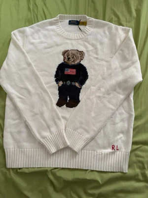 Vit stickad ralphlauren tröja med björnmotiv - Vit stickad tröja från Ralph Lauren med ikoniskt björnmotiv på bröstet, där björnen bär en tröja med amerikanska flaggan. Tröjan har rund hals, ribbstickade muddar och RL-broderi nertill. Det står Storlek L på den men den passar som en M eller S