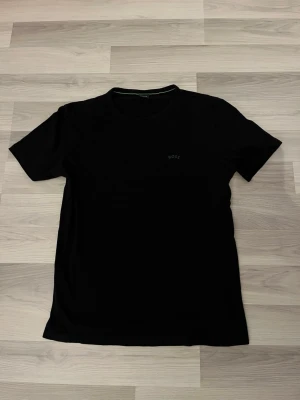 Svart t-shirt från BOSS - En stilren svart t-shirt från BOSS med diskret logga på bröstet. Klassisk passform och rund halsringning, perfekt till jeans eller shorts. Tillverkad i mjuk bomull för skön känsla hela dagen.