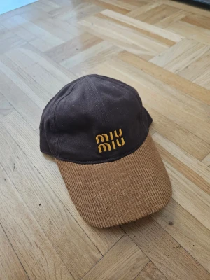 Brun keps från Miu Miu med brodyr - Snygg brun keps från Miu Miu med gul broderad logga framtill. Skärmen är i brunt manchestertyg och själva kepsen är i slät bomull. Perfekt accessoar för en trendig och avslappnad stil.
