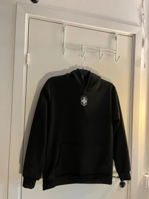 Svart hoodie med Brasilien-emblem - Säljer en svart hoodie med huva och stor magficka. På bröstet finns ett litet Brasilien-emblem i grönt, gult och vitt. Hoodien har ribbade muddar vid ärmar och nederkant. Perfekt för dig som gillar enkel stil med sportig touch.