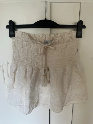 Vit kort kjol från zara med smock - Supersöt vit kjol från ZARA i storlek XS. Kjolen har smock i midjan, knytband med tofsar och ett lätt, luftigt tyg med diskret struktur. Perfekt för sommardagar och enkel att matcha med olika toppar.