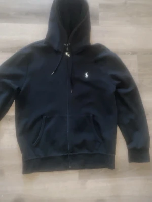 Svart hoodie med dragkedja från Polo Ralph Lauren - Säljer en svart hoodie från Polo Ralph Lauren med dragkedja framtill och klassisk huva med snörning. Tröjan har fickor på magen och den ikoniska lilla loggan broderad på bröstet. Perfekt för en avslappnad och stilren look.