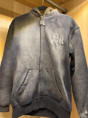 Grå zip hoodie från decrease  - Säljer en grå zip hoodie med huva och fickor framtill. På bröstet finns ett vitt tryck med texten 'DRECS' i gotisk stil och på huvan finns ett diskret broderat mönster. Hoodien har ribbade muddar och är tillverkad i mjukt bomullsmaterial och har använts sällan.
