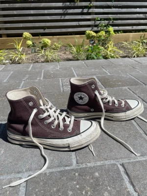Converse Chuck Taylor bruna highs - Klassiska Converse Chuck Taylor All Star i brunt canvasmaterial med vita gummisulor och tåhätta. Skorna har högt skaft, vita snören och den ikoniska loggan på sidan. Perfekta för en avslappnad och cool stil.