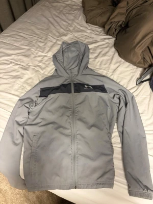 Under armour jacka - Ljusgrå vindjacka med dragkedja och huva. Jackan har en mörkgrå panel över bröstet och en liten logga på vänster sida. Tillverkad i lätt och vindtätt material, perfekt för blåsiga dagar. Två sidofickor och elastiska muddar vid ärmsluten.