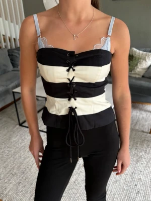 Randig korsettopp med snörning - Snygg korsettopp med breda svarta och vita ränder, justerbara ljusblå axelband och snörning framtill. Toppen har en figurnära passform och ger en edgy vibe till din outfit. Perfekt för dig som vill sticka ut med en unik topp.