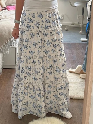 Vit långkjol med blå blommor - Romantisk långkjol i vitt med blått blommönster. Kjolen har flera volanger och en luftig passform som ger en bohemisk känsla. Perfekt för varma dagar och enkel att matcha med en enkel topp.