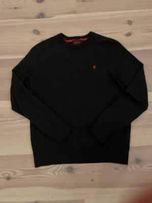 Svart stickad tröja från Morris - Snygg mörk grå stickad tröja från Morris i 100% merinoull. Tröjan har rund halsringning, långa ärmar och en liten orange logga broderad på bröstet. Perfekt för dig som gillar stilrena och klassiska plagg med en twist. Priset är förhandlingsbart 