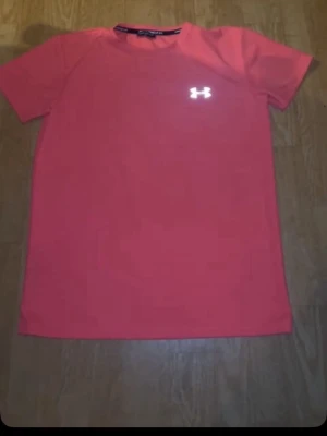 Rosa t-shirt från Under Armour - Snygg rosa t-shirt från Under Armour med klassisk logga på bröstet. T-shirten har korta ärmar och är tillverkad i ett mjukt och funktionellt material som passar perfekt till träning eller vardag.