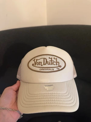 Beige Von Dutch truckerkeps - Snygg beige truckerkeps från Von Dutch med brunt broderat logotyp framtill. Kepsen har mesh-bakstycke för extra ventilation och justerbar snapback. Perfekt accessoar för en avslappnad streetwear-look. (Tar emot pris förslag)
