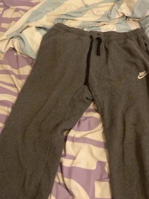 Gråa mjukisbyxor från Nike - Säljer ett par gråa mjukisbyxor från Nike med dragsko i midjan och resår. Byxorna har en klassisk passform och är tillverkade i mjukt sweatshirtmaterial. Perfekta för chill dagar eller när du vill vara bekväm.