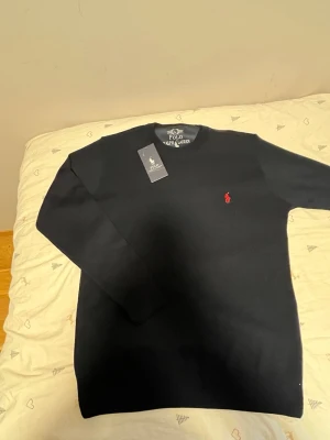 Svart tröja från Polo Ralph Lauren - Klassisk svart tröja från Polo Ralph Lauren med rund halsringning och den ikoniska röda logotypen broderad på bröstet. Tröjan har en enkel och stilren design, perfekt för dig som gillar tidlös stil. Materialet ser ut att vara mjukt och bekvämt.