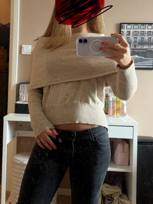 Beige off shoulder stickad tröja - Trendig beige stickad tröja med off shoulder-design och bred krage som viks ner över axlarna. Tröjan har långa ärmar och ribbade muddar vid ärmslut och nederkant. Perfekt för dig som gillar en stilren och modern look.