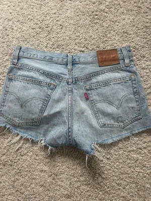 Ljusblåa jeansshorts Levi’s - Säljer ett par ljusblåa jeansshorts med råa, fransiga kanter och slitningar framtill. Shortsen har klassisk femficksdesign, knappgylf och en liten röd etikett bak. Perfekta för sommaren och ger en avslappnad vibe.