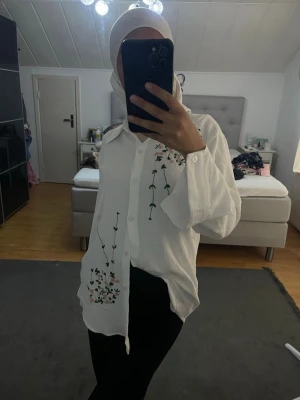Vit skjorta med blommigt broderi - Säljer en vit skjorta med blommigt broderi i grönt, svart och orange. Skjortan har lång ärm, klassisk krage och knappar framtill. Passformen är loose och materialet känns som bomull. Perfekt för dig som gillar unika detaljer! Aldig använd 