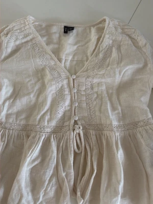Krämvit boho-blus med spetsdetaljer - Söt krämvit blus i boho-stil med dekorativa spetsdetaljer längs v-ringningen och knappar framtill. Blusen har ett knytband i midjan och är tillverkad i ett tunt, luftigt tyg som känns som bomull eller linne. Perfekt för en avslappnad och trendig look. Den e då lite sönder vid slutet på armarna men är inget man ser ( går o sy) köpt för 600